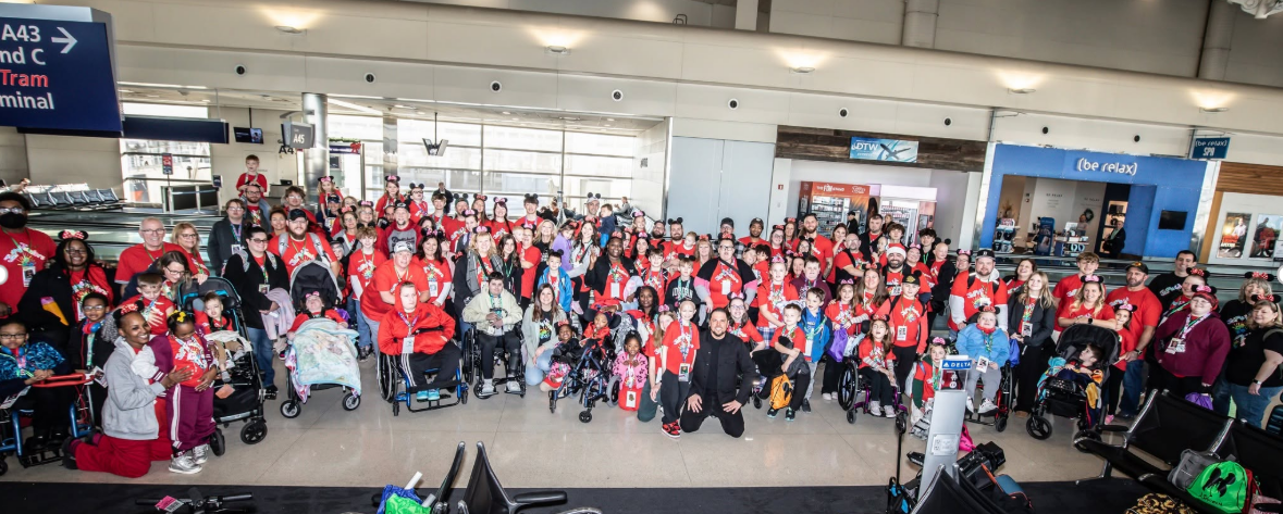 Large diverse group of people in an airport terminal posing for a group photo, many wearing red shirts and some in wheelchairs or strollers.