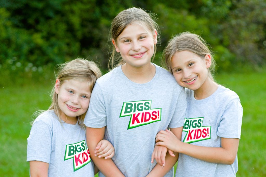 Three smiling children in gray shirts with a green-and-red 'BIGS KIDS!' logo, standing arm-in-arm in a grassy outdoor setting.