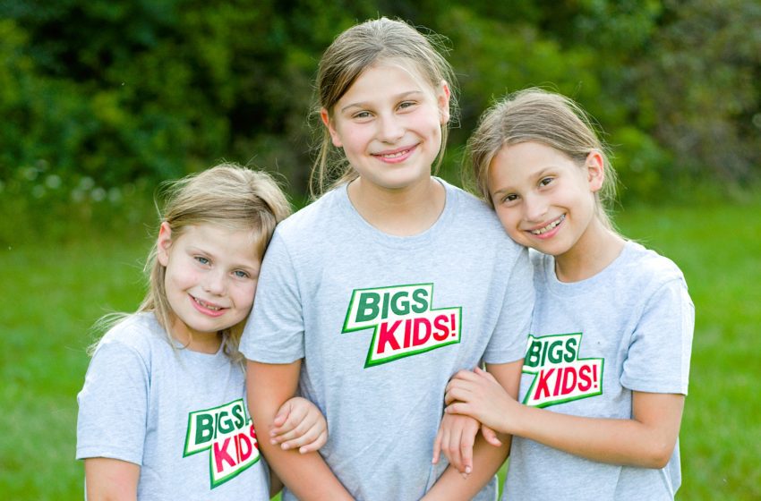Three smiling children in gray shirts with a green-and-red 'BIGS KIDS!' logo, standing arm-in-arm in a grassy outdoor setting.