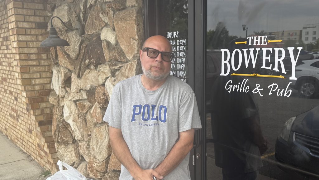 Man wearing a gray 'Polo Ralph Lauren' t-shirt stands outside a storefront with a stone wall and a glass door that reads 'The Bowery Grille & Pub'.