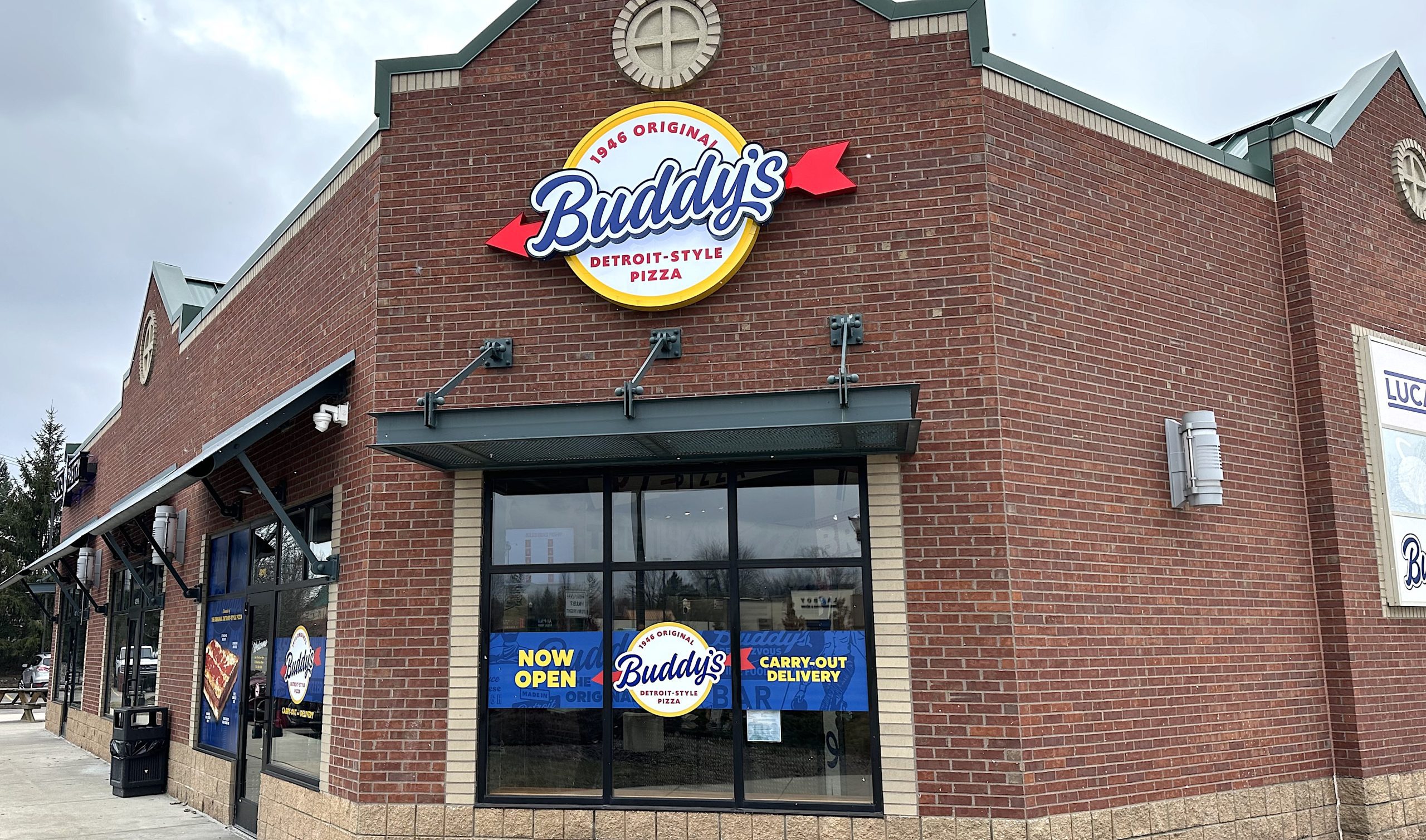 Exterior view of Buddy's Detroit-Style Pizza restaurant on a brick building with a large circular yellow logo sign reading 'Buddy's Detroit-Style Pizza'.