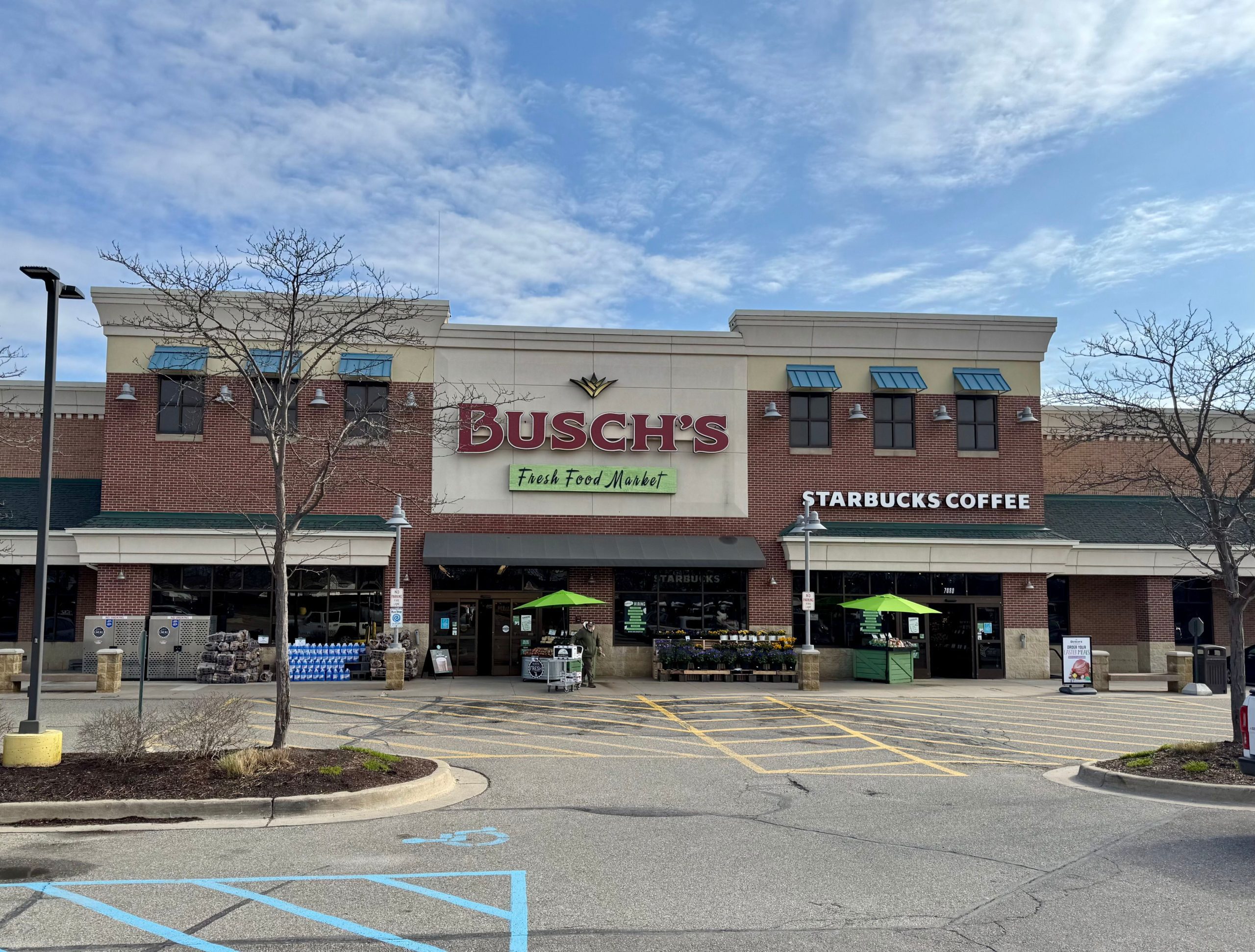 Busch's Fresh Food Market storefront in a strip mall with a Starbucks Coffee sign, under a blue sky with thin clouds and leafless trees in front.
