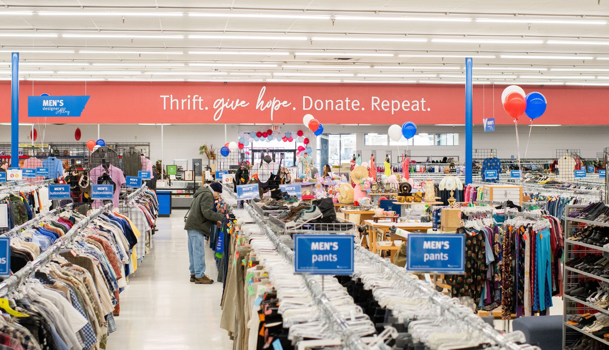 Inside a busy thrift store with clothing racks and balloons, and a red banner reading 'Thrift. give hope. Donate. Repeat.'