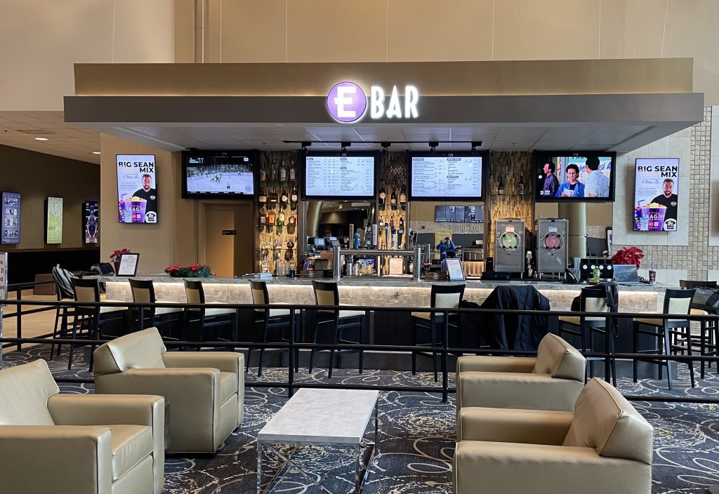 Interior of a modern hotel lobby bar with a long dark counter, beige seating, and digital menu screens behind the bar; a purple circular 'E' logo above reads 'BAR'.