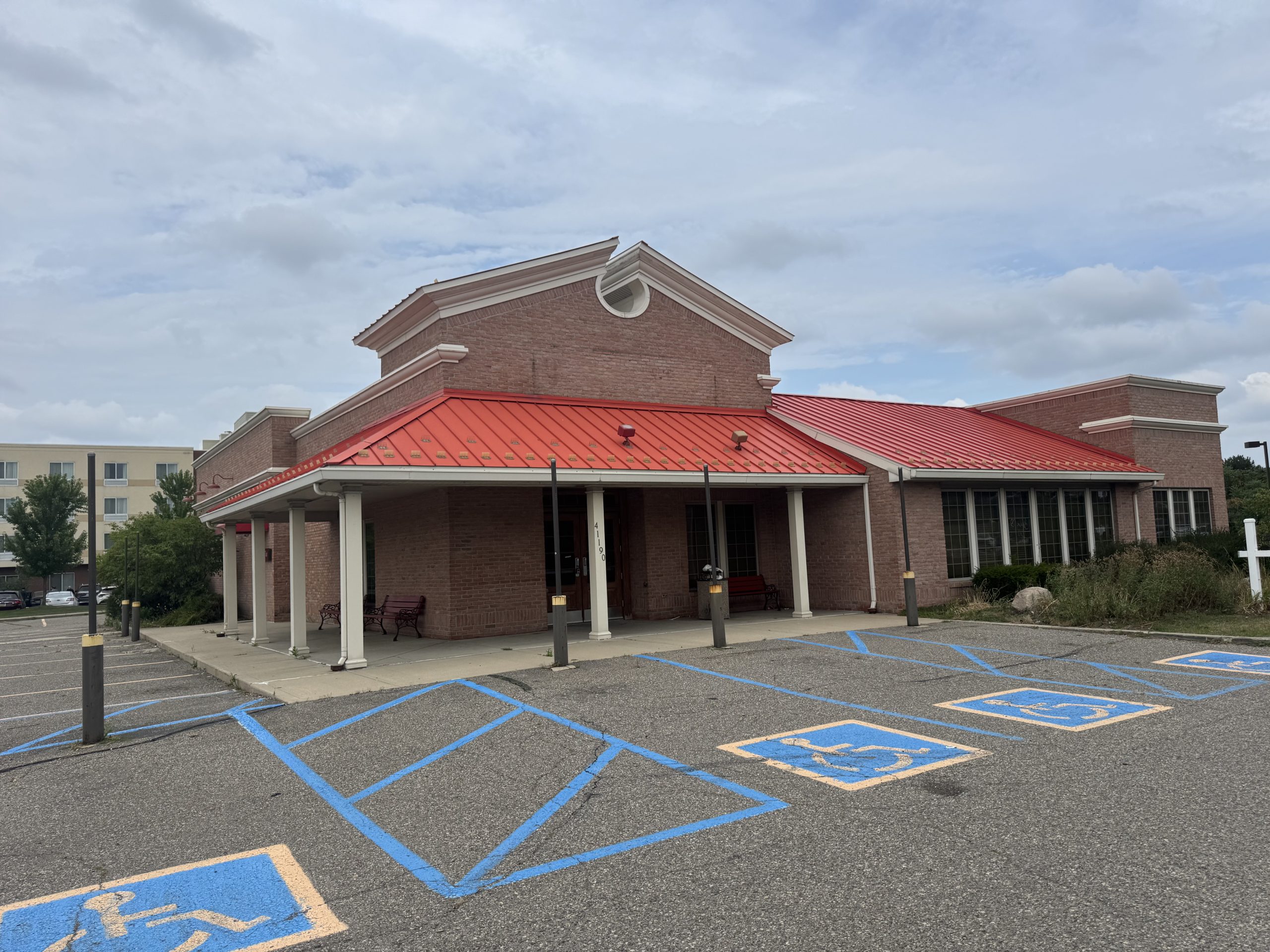EvansChik - Social House News Brick building with a red metal roof and covered entrance against a cloudy sky; accessible parking stalls marked in blue in the foreground.