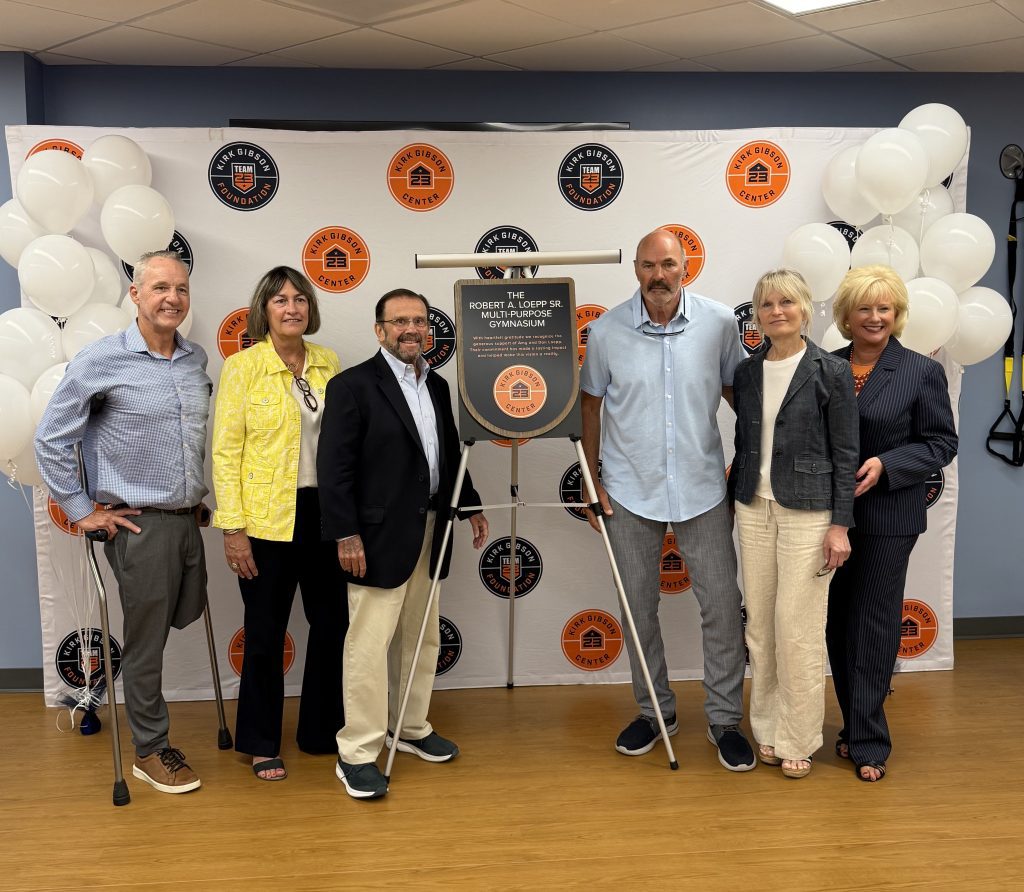 Six adults pose for a photo in front of a branded backdrop with orange and black circular logos and white balloons on both sides, indoors.