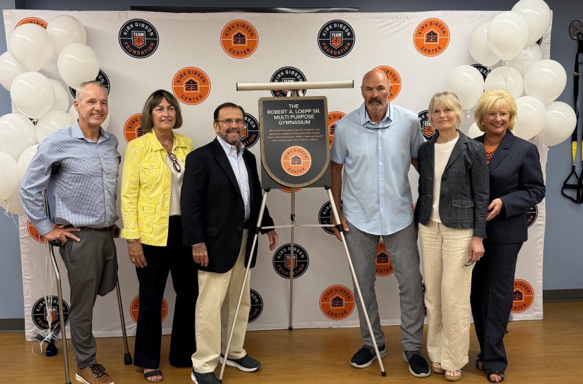 Six adults pose for a photo in front of a branded backdrop with orange and black circular logos and white balloons on both sides, indoors.