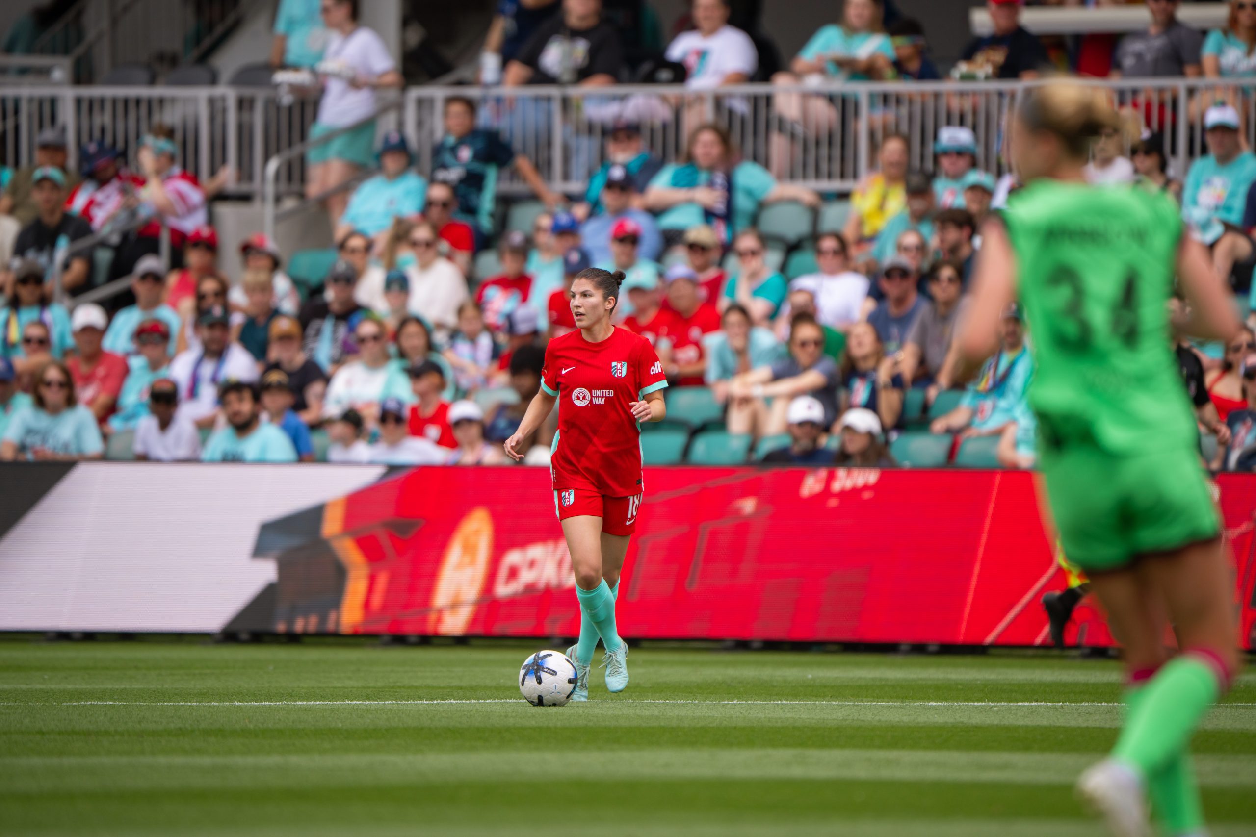 Izzy1 - Social House News Female soccer player in red runs with the ball on the green field as a crowd watches from the stands.