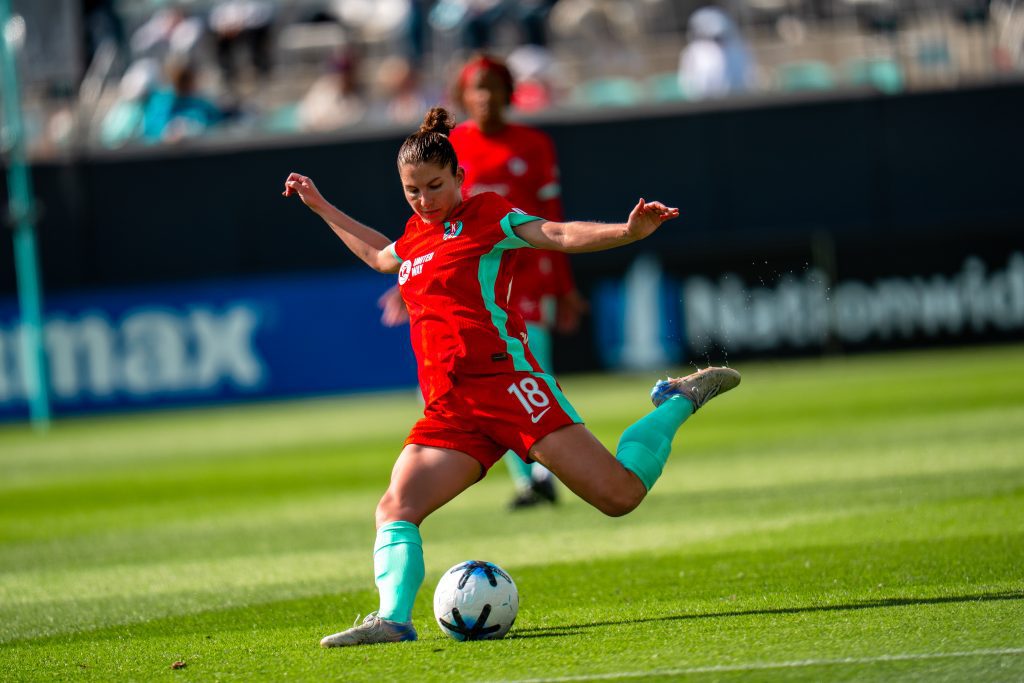 Soccer player in a red kit with turquoise socks kicking a ball on a sunny, green field.