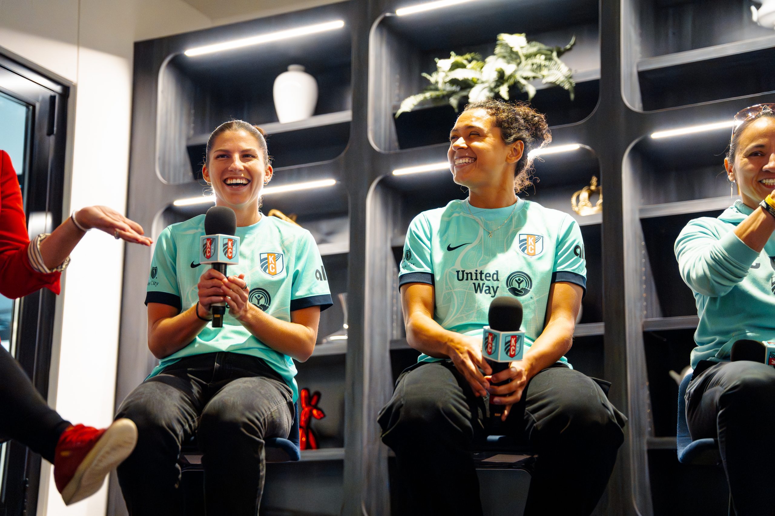 Izzy3 Sideline the Stigma00134 - Social House News Three women in teal KC jerseys sit on a stage, laughing and holding microphones during an interview.