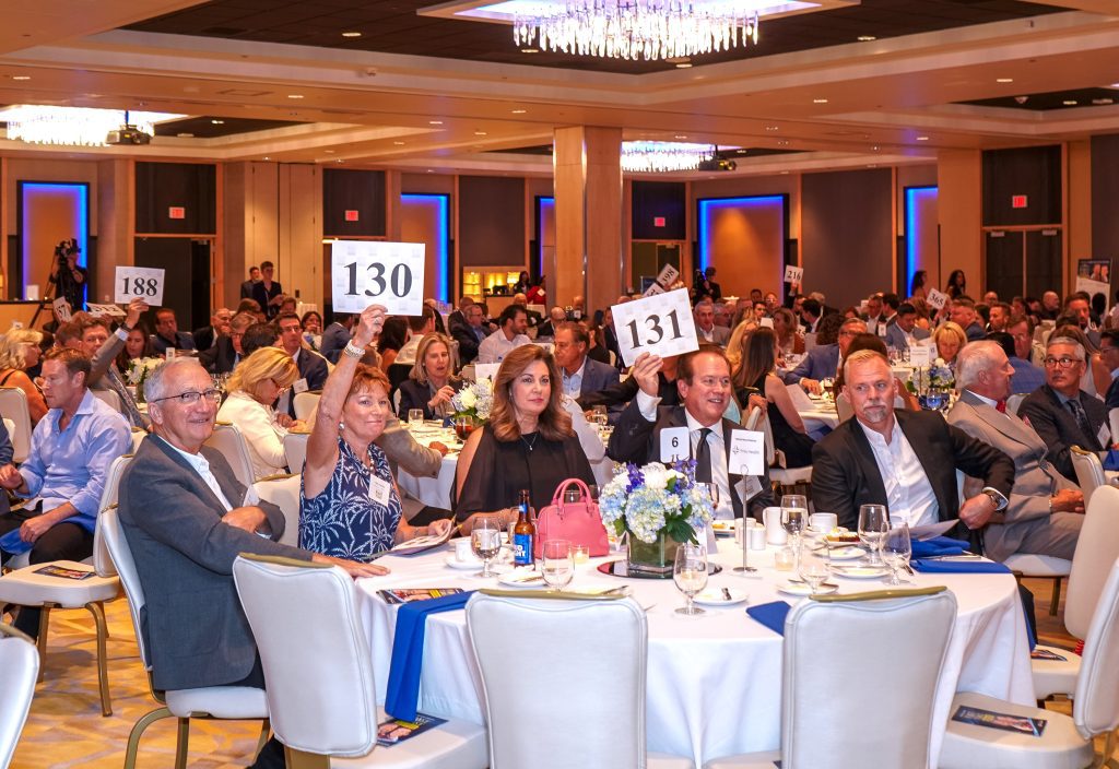 Crowded banquet hall during a formal gala; guests raise numbered signs (130, 131, 188) at their tables.