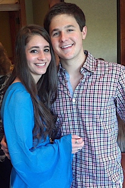 Two smiling young adults posing indoors, a woman in a bright blue top and a man in a red-and-white checkered shirt, standing close together with arms around each other.