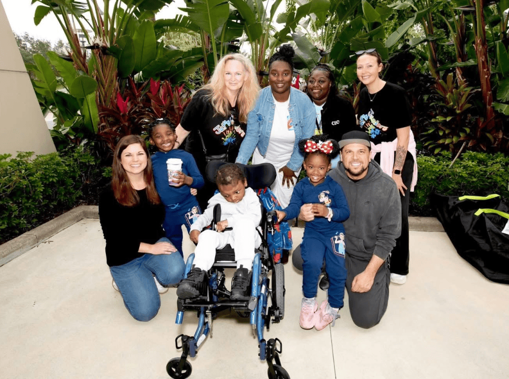 Group of adults and children posing outdoors among tropical plants, smiling at the camera with a child in a wheelchair in the center.