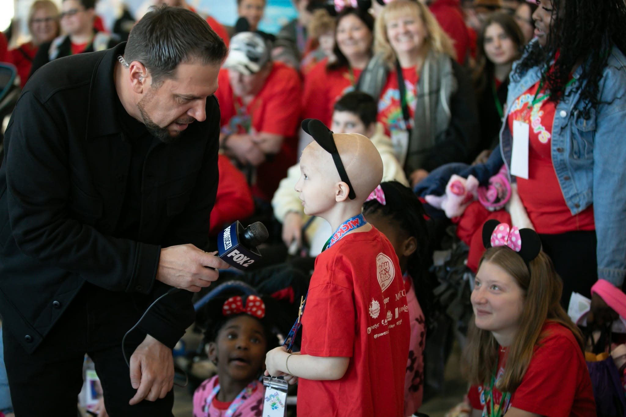 Reporter kneels to interview a young girl in a red shirt, microphone labeled FOX2, as a crowd in red shirts watches.