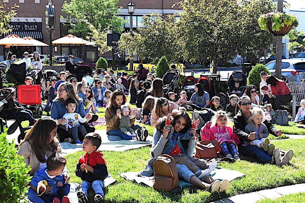 Large group of families sitting on blankets in a sunny park for an outdoor community event, with kids and strollers nearby.