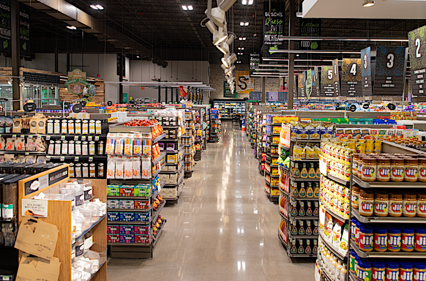 Wide view of a busy grocery store aisle with tall shelves stocked with jars, boxes, and beverages under bright ceiling lights.
