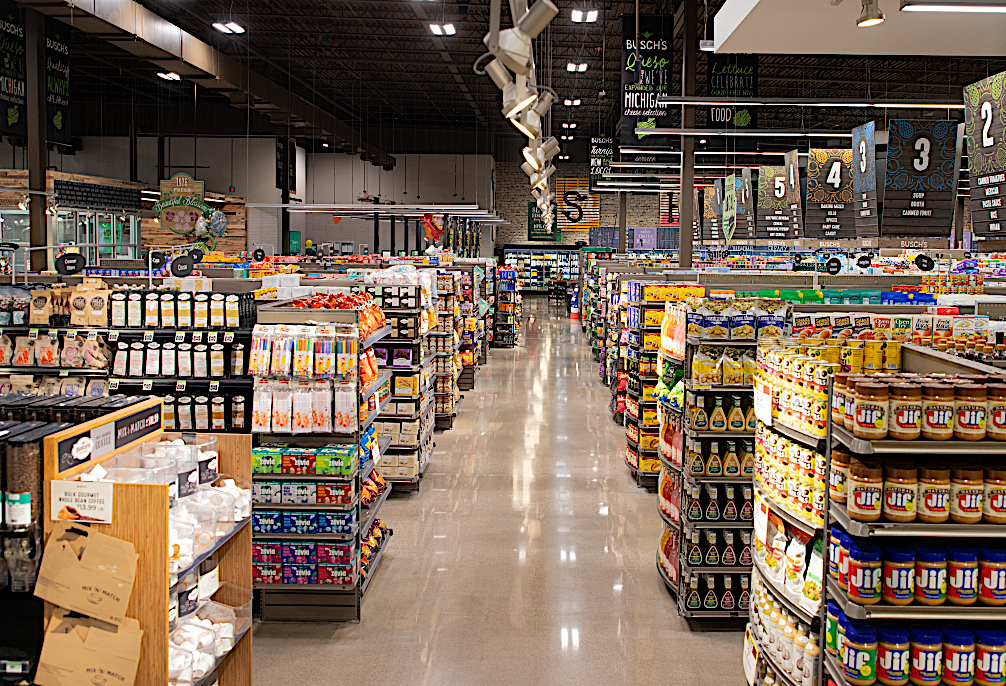 Wide view of a busy grocery store aisle with tall shelves stocked with jars, boxes, and beverages under bright ceiling lights.