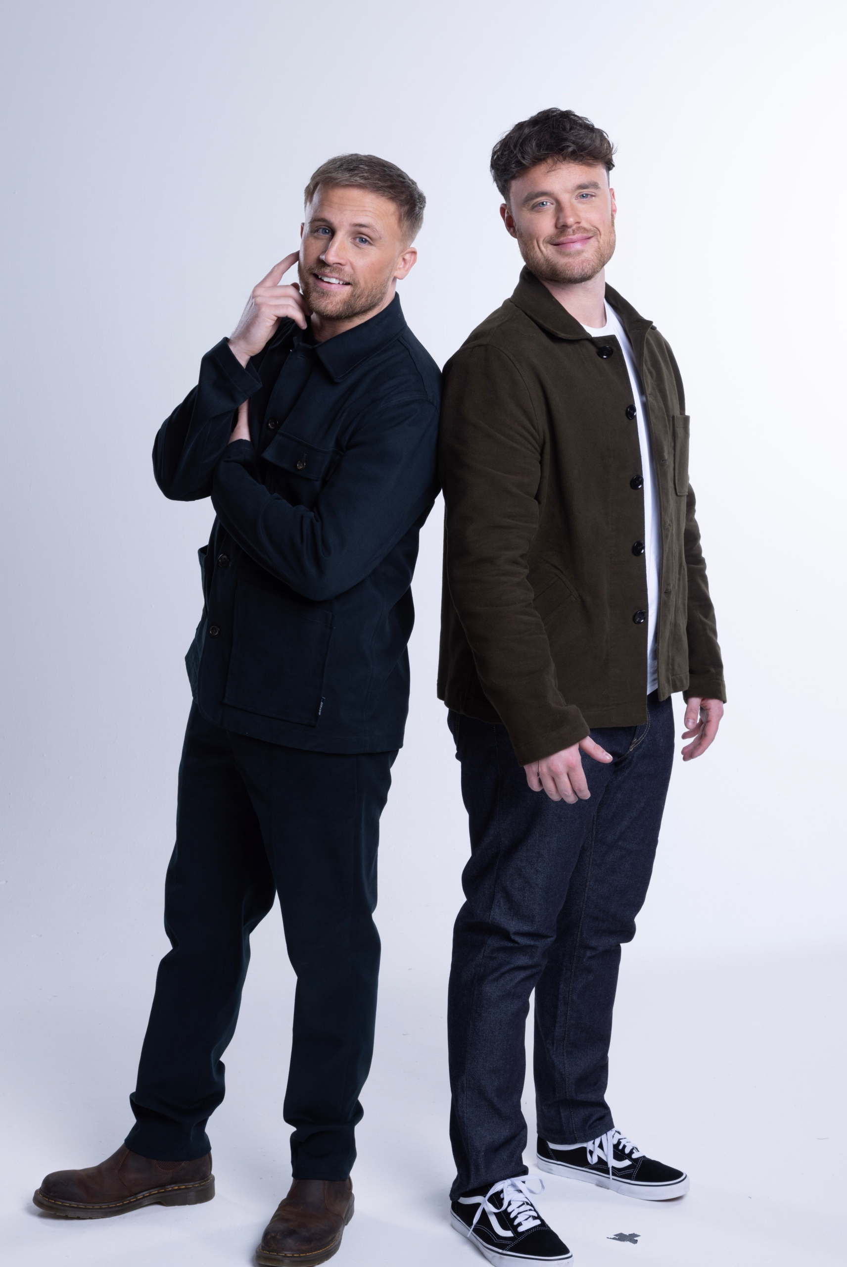 Two men stand back-to-back, smiling at the camera in a studio; left man in a dark navy jacket with brown boots, right man in an olive jacket with jeans and black-and-white sneakers.