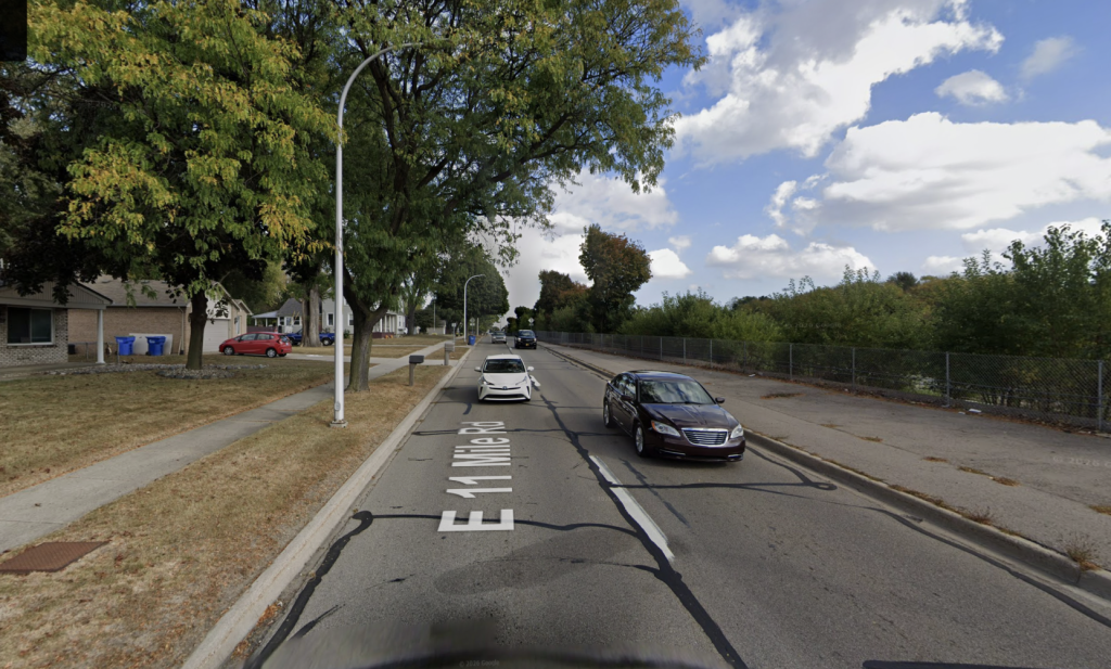 Two-lane suburban street with cars driving and parked along the curb, trees lining the sidewalks under a blue sky.