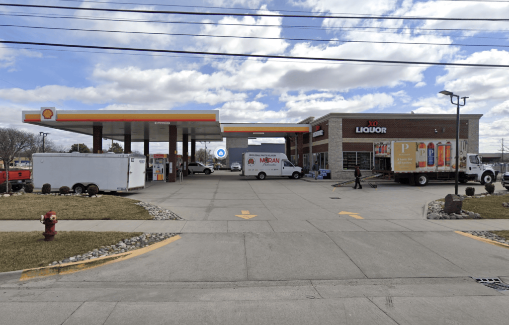 Shell gas station with a convenience store labeled XO Liquor on the right; delivery vans and a white truck are in the forecourt under a blue sky with scattered clouds.