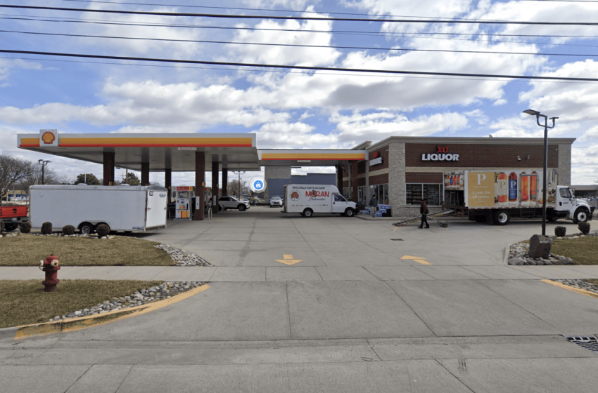 Shell gas station with a convenience store labeled XO Liquor on the right; delivery vans and a white truck are in the forecourt under a blue sky with scattered clouds.