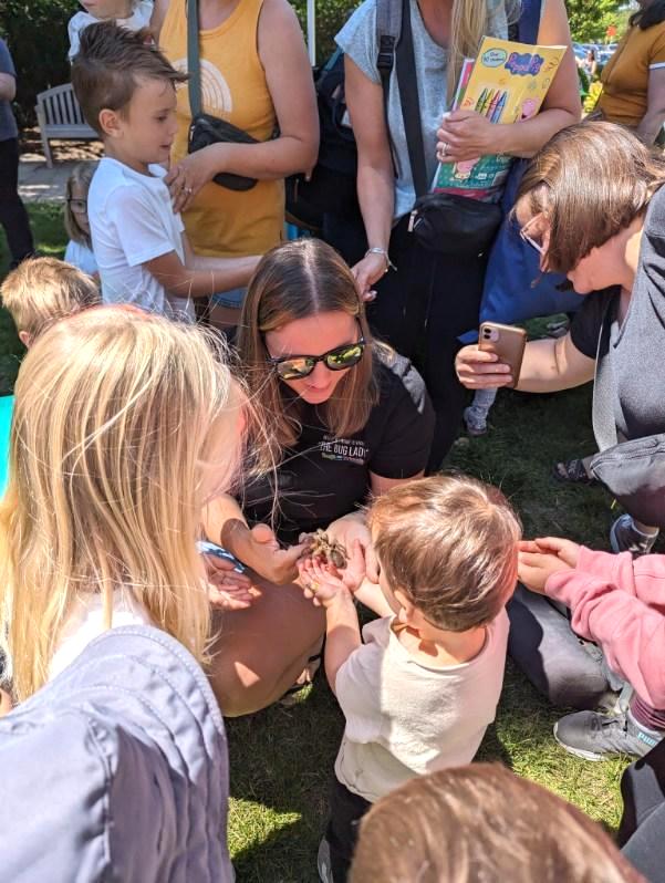 VORH_BugonWheels4 - Social House News Woman kneeling on grass showing a small animal to several children at an outdoor event in a park.