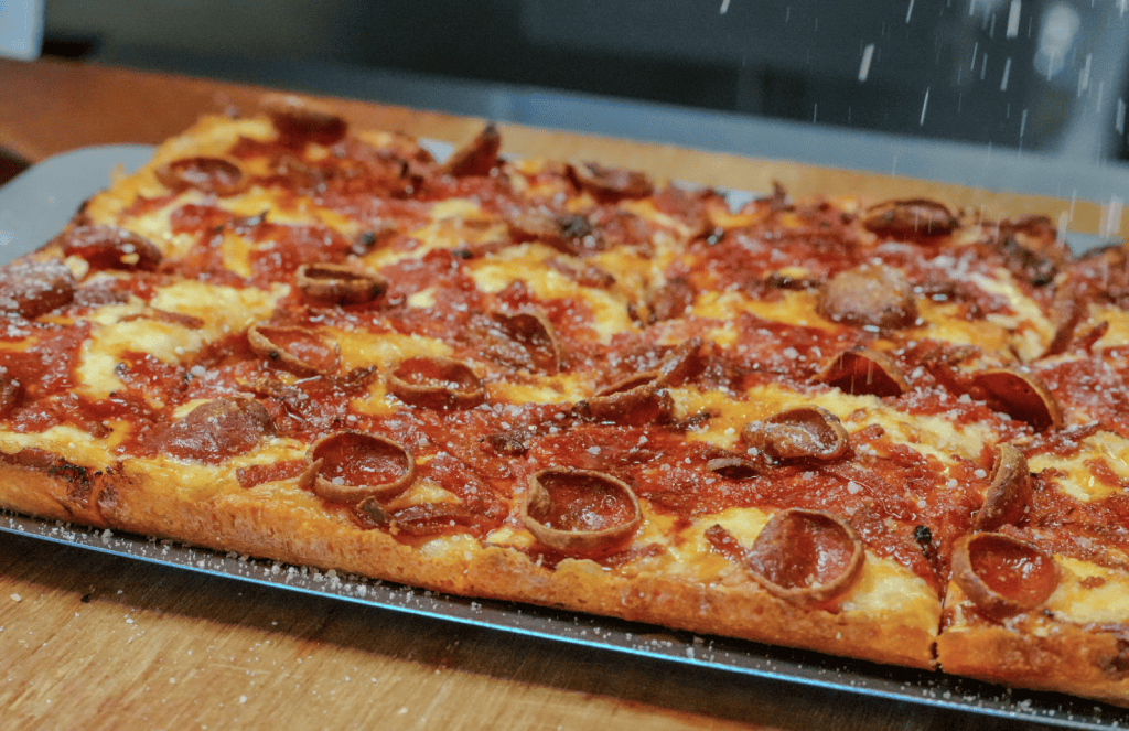 Rectangular pepperoni pizza on a metal baking sheet with melted cheese and pepperoni slices, resting on a wooden counter