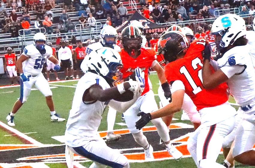 Football game action: a white-team defender reaches for an orange-team ball carrier as others converge on the field with a crowded stadium in the background.