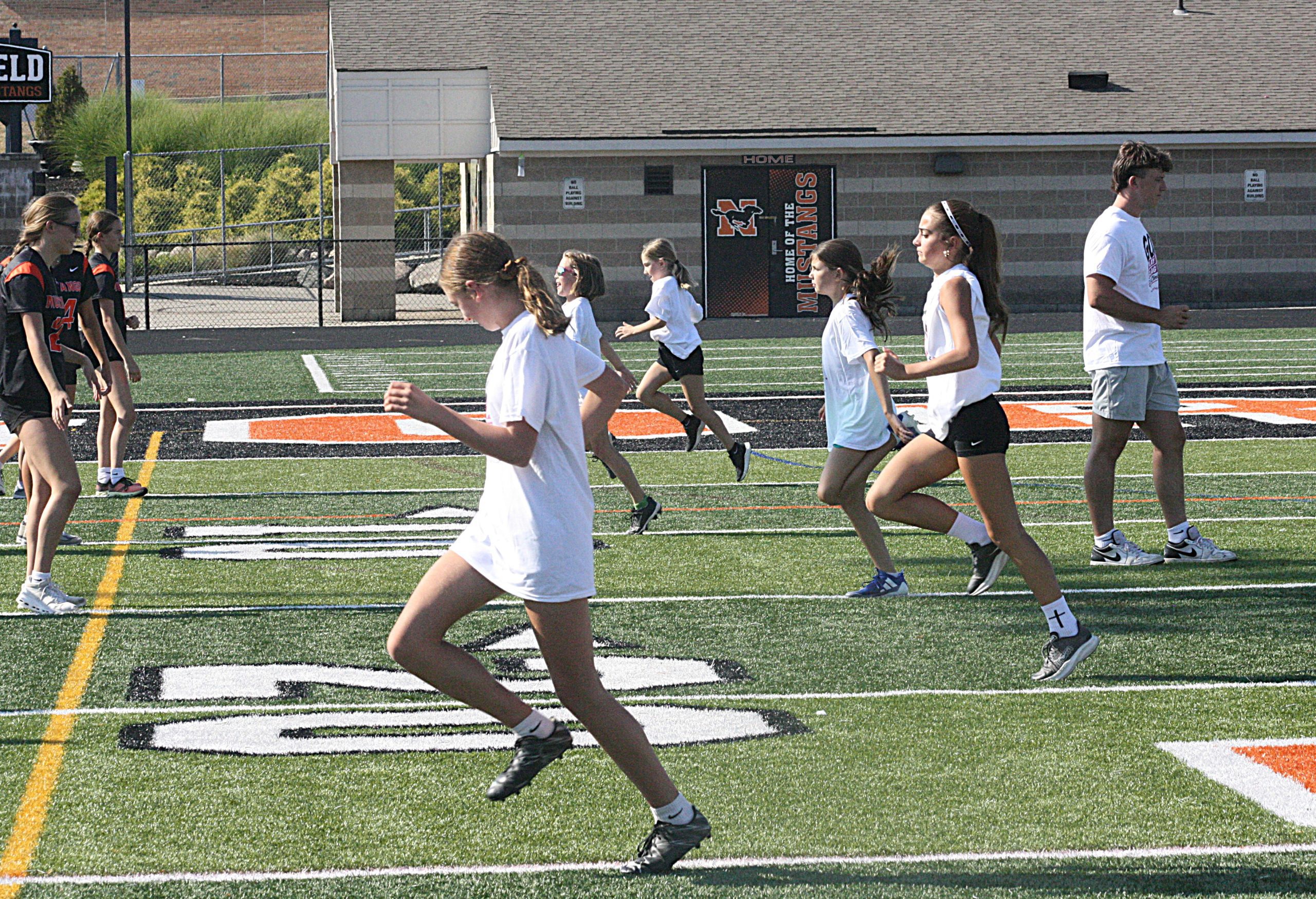 girlsff3 1 - Social House News Group of young female runners in white shirts sprint on a football field with a school stadium in the background.