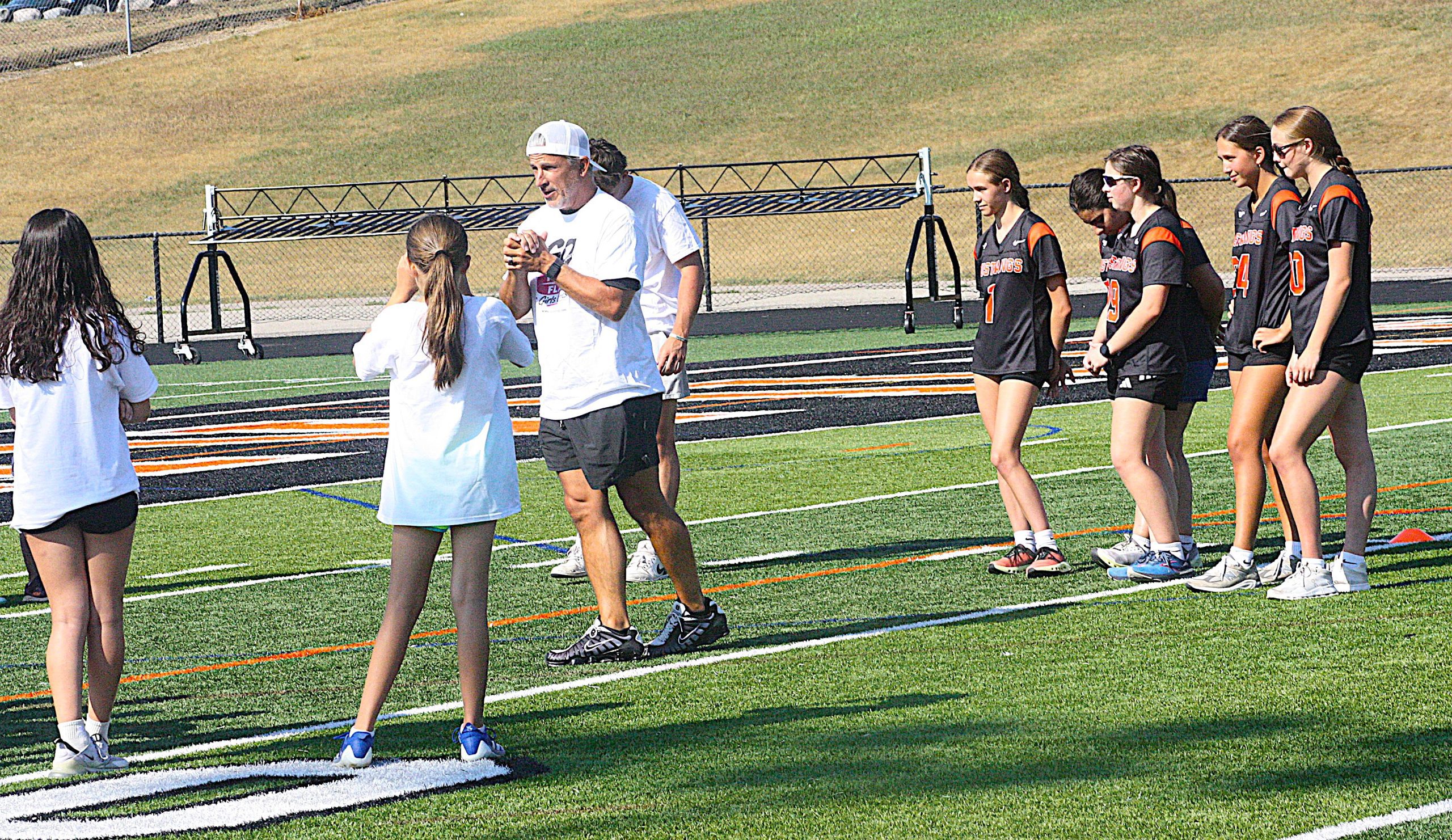 Line of female athletes in black and orange uniforms on a track field, coach in white shirt high-fiving a player in white at center.