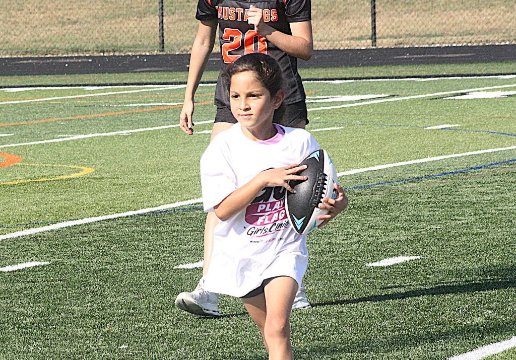 Young girl in a white shirt runs on a synthetic football field while holding a football, with a taller player in black behind her in the background.