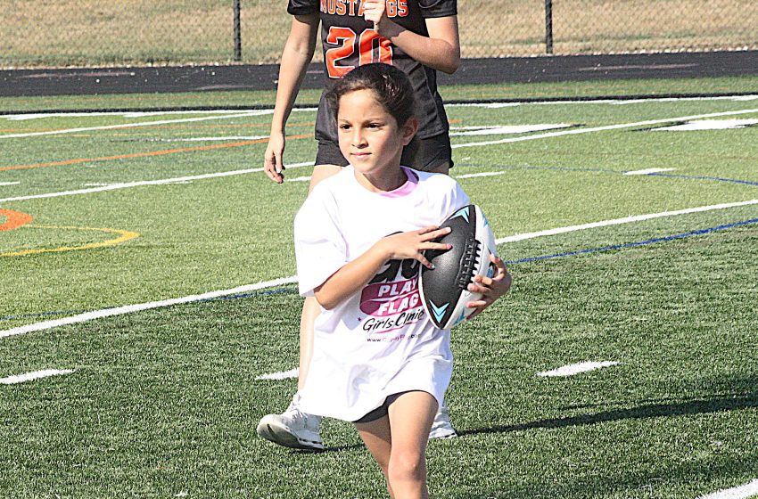 Young girl in a white shirt runs on a synthetic football field while holding a football, with a taller player in black behind her in the background.